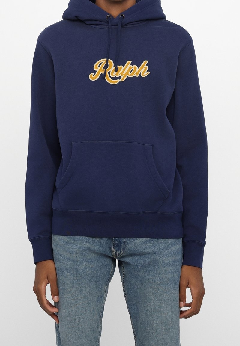 Person som bär en marinblå huvtröja med gula "Ralph"-bokstäver och ljusblå denimjeans, stående mot en neutral bakgrund.