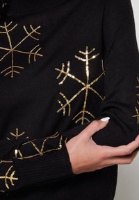 Schwarzer Pullover mit goldenen Pailletten-Schneeflocken-Designs an den Ärmeln. Das Material ist gestrickt, mit gerippten Bündchen und einer glatten Textur.