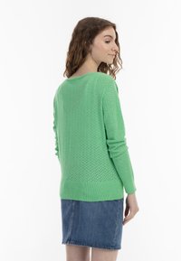 Maglione verde lavorato a maglia con un motivo testurizzato, scollo rotondo e polsini a coste, abbinato a una gonna di jeans. La vista frontale evidenzia il retro testurizzato.