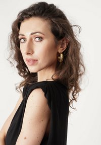 Czarna sukienka z tkaniny, z falbanami na ramionach, o miękkiej teksturze. Modelka ma kręcone ciemne włosy i złote kolczyki w kształcie kół. Skóra ma lekkie piegi.