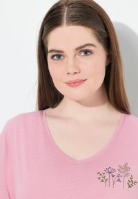 Růžové tričko s V výstřihem, které má květinový vzor v tmavě zelené a černé výšivce na levé straně hrudi. Měkká, lehká textilie.