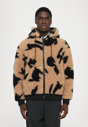Man wearing a tan and black patterned fleece zip-up hoodie with a hood and black pants, standing against a plain background.
