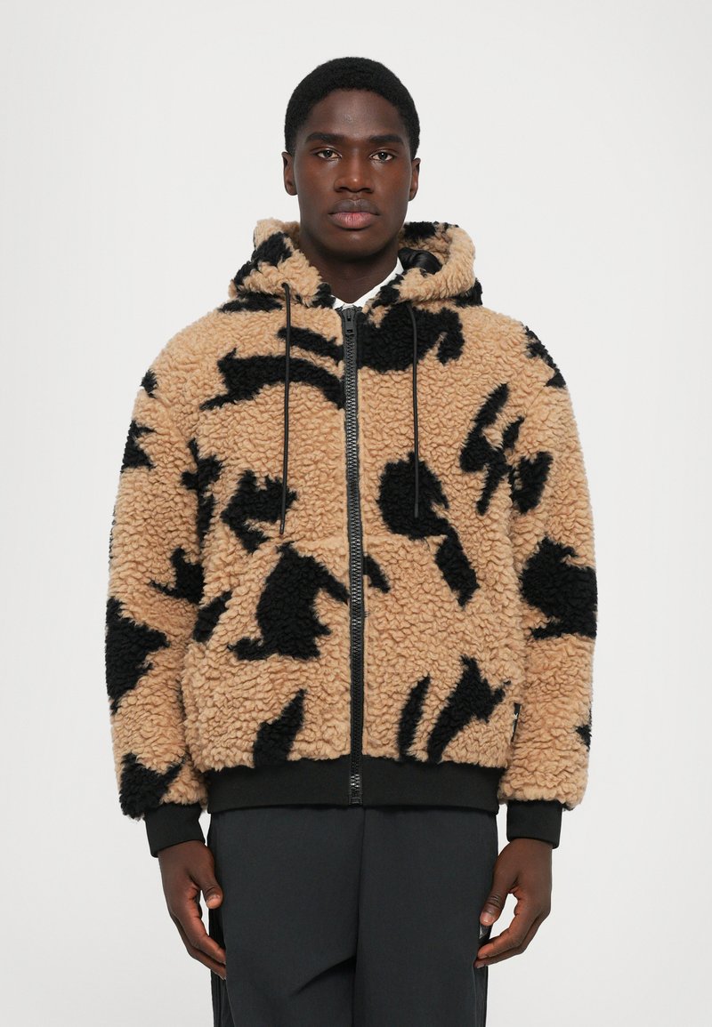 Man in een beige en zwarte gemusterde fleece zip-up hoodie met een capuchon en zwarte broek, staande tegen een effen achtergrond.