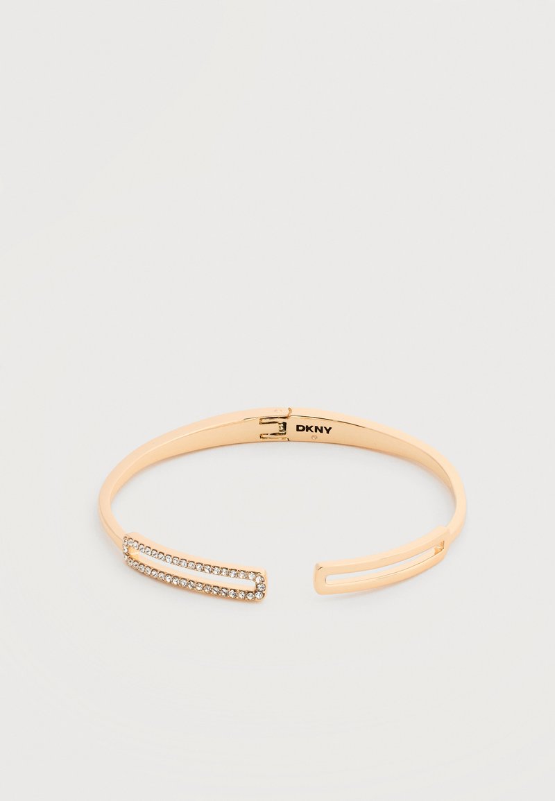 Ανοικτό βραχιόλι σε χρυσό τόνο με ορθογώνιες κρυστάλλινες λεπτομέρειες στη μία άκρη και λογότυπο DKNY στο εσωτερικό.