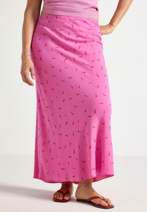 Donna che indossa una gonna lunga rosa con piccolo motivo di peperoncini rossi, un top rosa, sandali rossi e un braccialetto dorato alla caviglia, in piedi su sfondo bianco.
