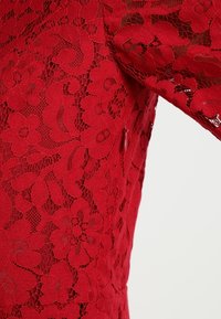 Punainen pitsimekko, jossa on kukkakuosiset yksityiskohdat, muotoiltu leikkaus ja näkyvä vetoketju sivussa. Tekstuuri vaikuttaa monimutkaiselta ja herkältä.