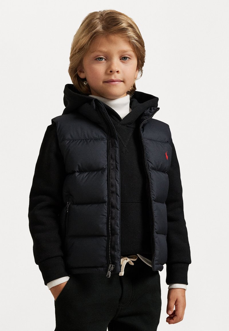 Colete acolchoado azul marinho com fecho de correr e bolsos laterais, sobreposto a um moletom preto. Suéter de gola alta branco por baixo, combinado com calças escuras.