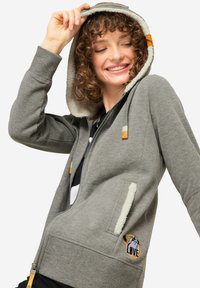 Felpa grigia con zip, cappuccio foderato in pile, texture morbida, tasche frontali e un'applicazione ricamata di un cane. Gli accessori presentano dettagli arancioni.