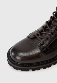 Stivale in pelle marrone scuro con suola nera robusta e lacci marroni abbinati, che mostra pelle strutturata e cuciture rinforzate su sfondo bianco.