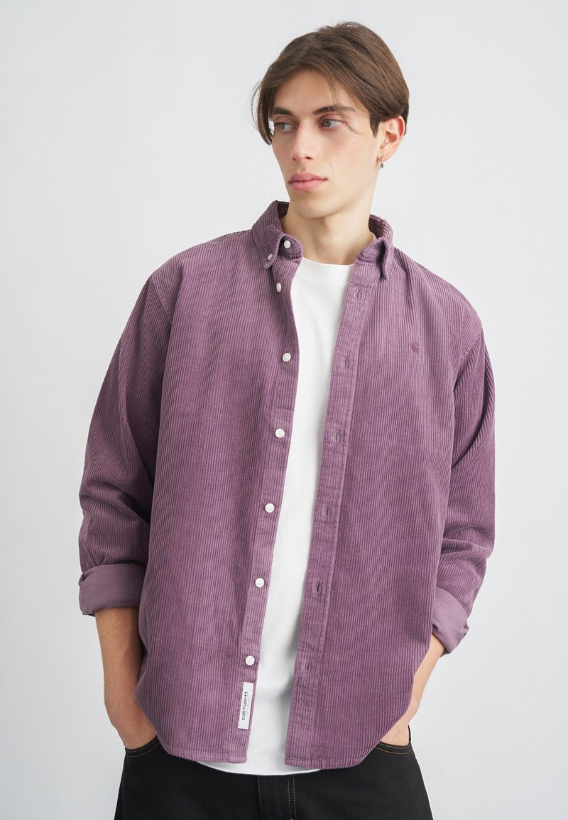 Dlhý rukáv košele z jemnej kordurovej látky vo svetlo fialovej farbe s golierom na gombíky, bielymi gombíkmi a malým logom. Nosená cez biele tričko.