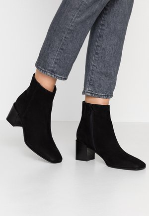 Czarne zamszowe botki za kostkę z szpiczastym noskiem, zamkiem błyskawicznym po boku i obcasem na słupku. Noszone z krótkimi szarymi jeansami.