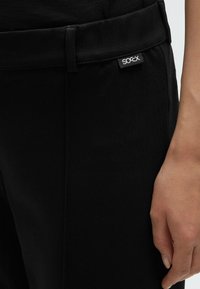 Black pants with a smooth texture, featuring a flat waistband and a small white logo tag. Close-up of hand positioned near the waistband.