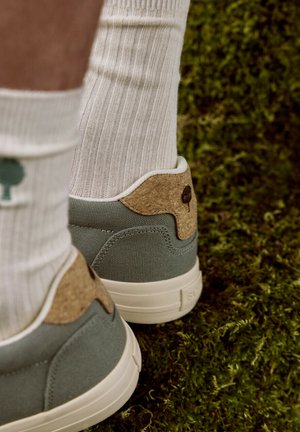 Pieds portant des chaussettes blanches côtelées avec des icônes d'arbres verts et des baskets vertes avec des accents en liège, debout sur de la mousse verte.