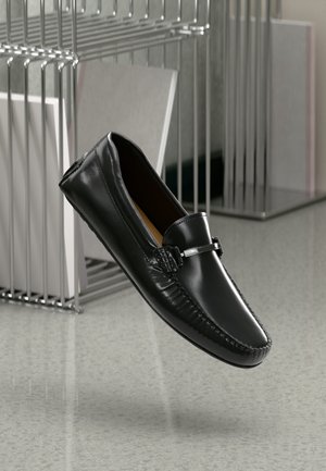 Zwarte leren loafer met een ronde neus, geprofileerde details en een metalen accent op de wreef, tegen een grijze achtergrond.
