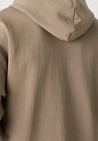 Bluza z kapturem wykonana z miękkiej beżowej tkaniny. Charakteryzuje się luźnym krojem, gładką teksturą i minimalistycznym designem, podkreślającym kontury kaptura.