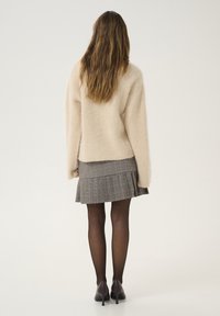 Beige fuzzy trui met lange mouwen, gecombineerd met een geplooide grijze geruite rok en zwarte panty, afgewerkt met donkere hakken.