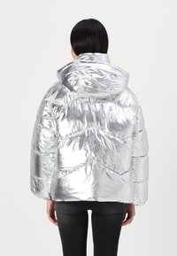 Chaqueta de plumón plateada con capucha, que presenta un acabado brillante, textura acolchada y patrón de chevrón. Cortada a media longitud con un ajuste Oversize.