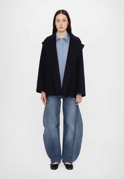 Cappotto in misto lana navy con ampi revers, camicia azzurro chiaro, e jeans in denim blu a gamba larga, abbinati a scarpe basse nere.
