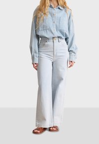 Camisa de mezclilla de gran tamaño en azul claro con dos bolsillos en el pecho, combinada con jeans de pierna ancha de cintura alta en azul claro y sandalias marrones.