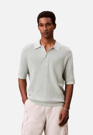 Hombre con polo de punto de manga corta color gris claro y pantalones cortos cargo beige, de pie con una mano en el bolsillo.