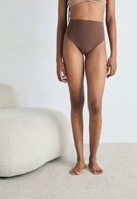 Brune høyhalsede undertøy med en glatt tekstur og minimalt design. Modellen står barføtt i en nøytral setting nær en buet, lyse farget stol.