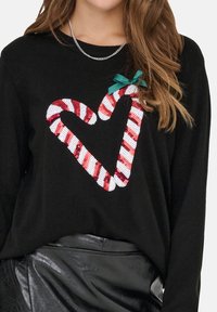 Schwarzer Pullover mit einem herzförmigen Design aus rot-weißen Zuckerstangenstreifen, verziert mit Pailletten und einem grünen Schleifenakzent.