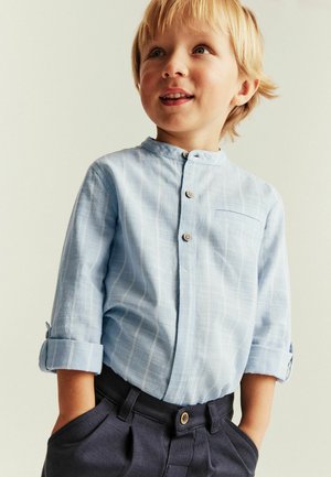 Blonder Junge trägt ein hellblaues Hemd mit Knöpfen und hochgekrempelten Ärmeln sowie dunkle Hosen, schaut leicht nach oben und lächelt sanft.