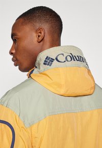 Gul og lys grøn vindjakke med mesh-detaljer, der har hætte og Columbia-branding på kraven. Letvægtig kvalitet.