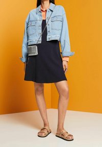 Kurtka dżinsowa typu cropped na czarnej sukience, widoczny pomarańczowy kołnierzyk. Beżowe sandały i mała srebrna torebka na ramię dopełniają outfit. Pomarańczowe tło.