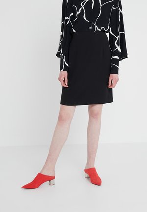 Jupe noire au design cintré, associée à un chemisier à motifs noir et blanc et à des mules en daim rouge avec des talons compensés métalliques.
