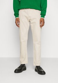 Beige Baumwollhosen mit einem geraden Bein, die einen schlichten Schnitt aufweisen. Kombiniert mit schwarzen Dressschuhen und einem grünen Strickpullover.