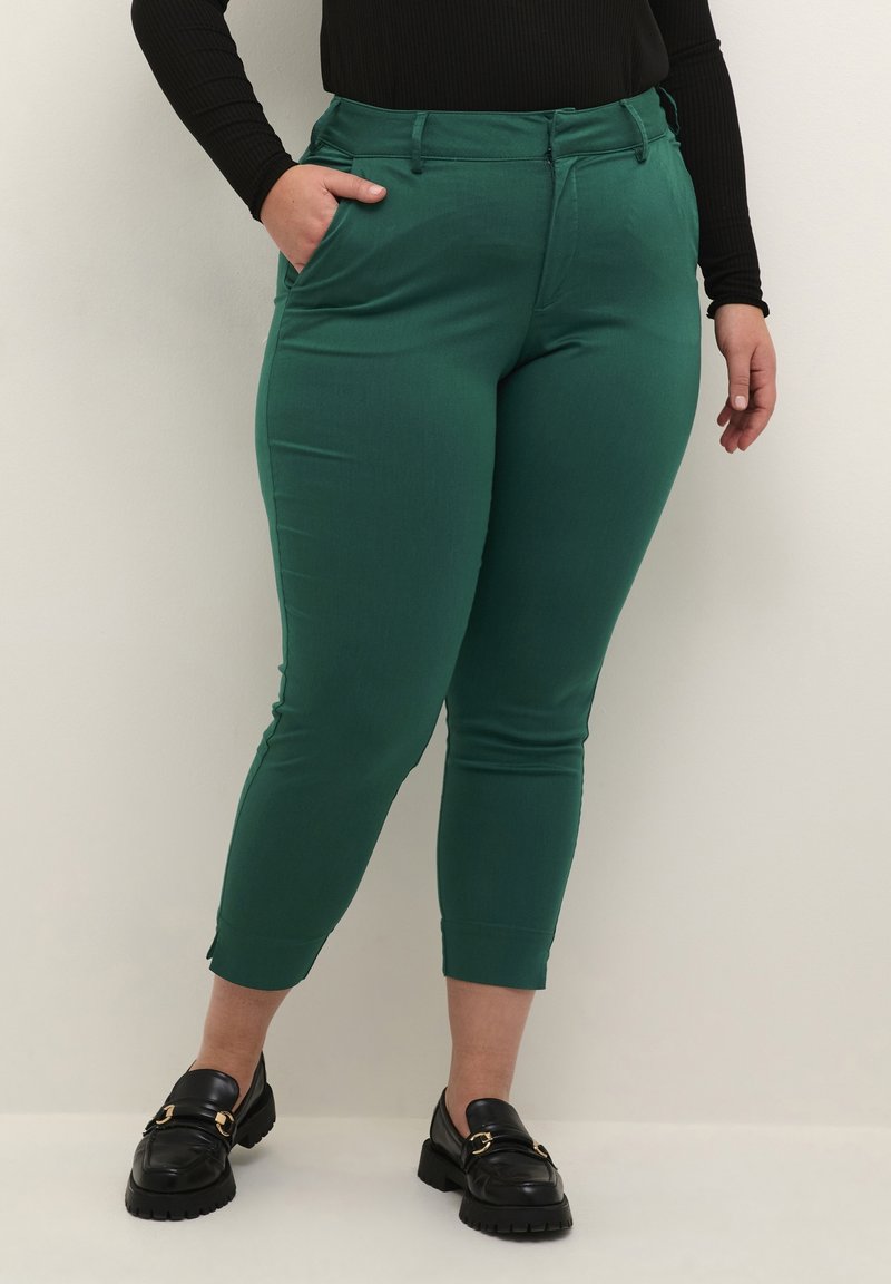 Vihreät, istuvat housut, jotka on valmistettu sileästä kankaasta, keskikorkealla vyötäröllä, sivutaskuilla ja kapenevalla nilkkaleikkauksella, yhdistettynä mustiin loafer-kenkiin.