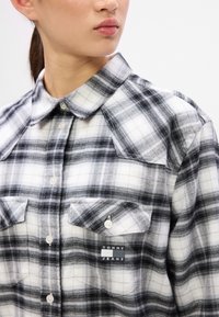 Camisa de cuadros en blanco y negro hecha de una tela suave, con un collar clásico, dos bolsillos frontales y una etiqueta de logo en el bolsillo izquierdo.