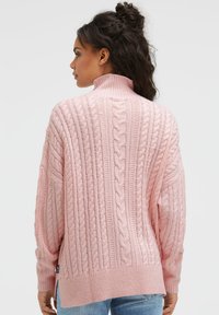 Hellrosa gestrickter Pullover mit hohem Kragen, mit Zopfmuster, langen Ärmeln und seitlichen Schlitzen am Saum.