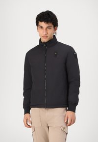 Black zip-up jacket with high collar, elastic cuffs, and two front pockets. Features a green accent on the left side and a patch on the shoulder.