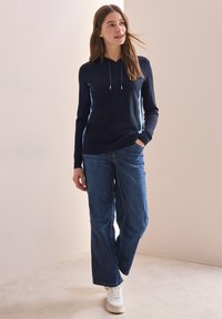 Navy Kapuzenpullover mit hellblauen seitlichen Streifen, kombiniert mit weiten blauen Jeans und weißen Sneakers. Glatte Textur, lässige Passform.