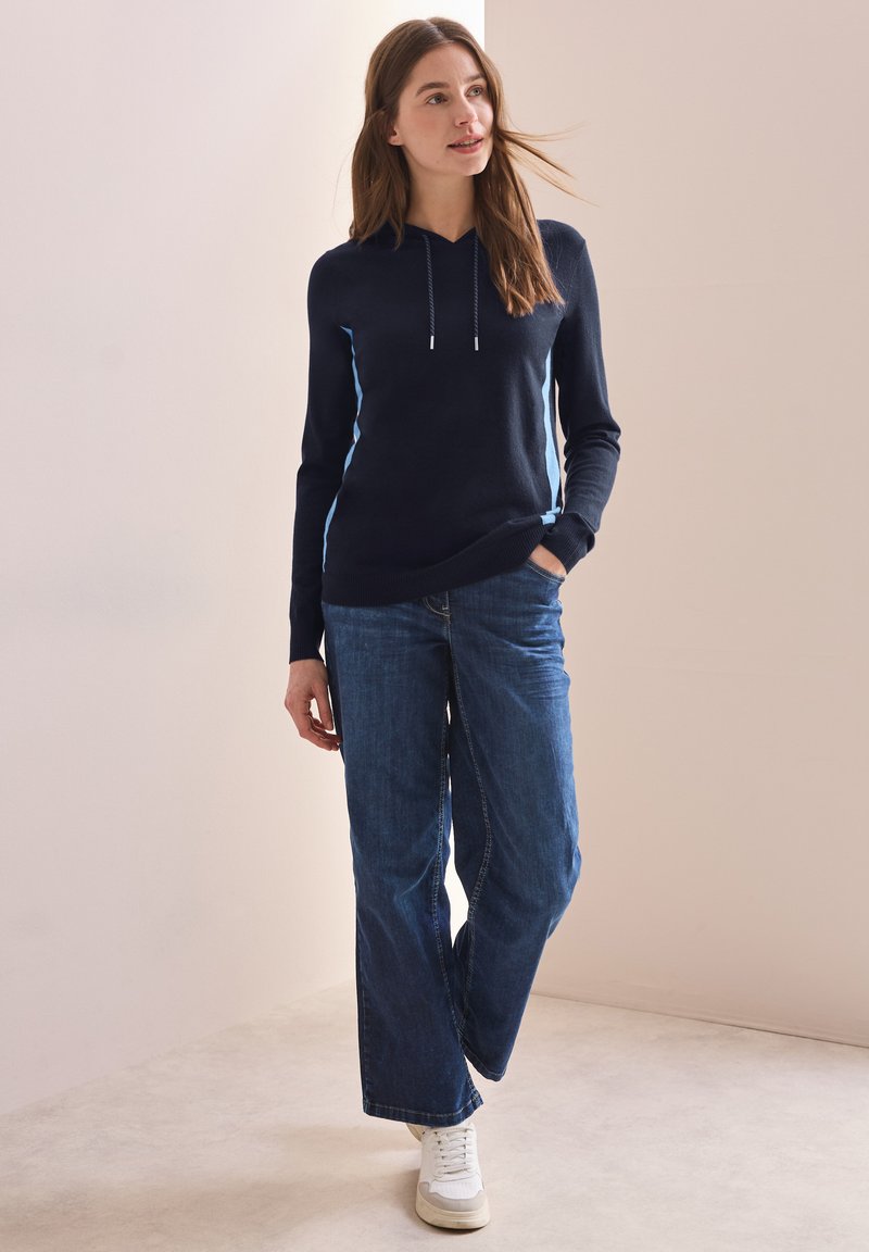 Navy Kapuzenpullover mit hellblauen seitlichen Streifen, kombiniert mit weiten blauen Jeans und weißen Sneakers. Glatte Textur, lässige Passform.