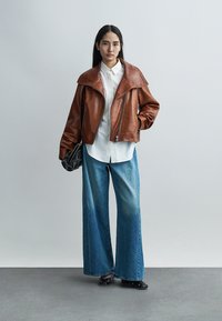 Young woman stands wearing a brown leather jacket, white button-up shirt, wide-leg blue jeans, black shoes, and holding a black quilted handbag.