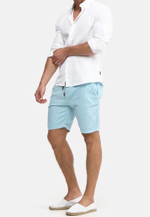 Camicia bianca in lino a maniche lunghe, pantaloncini azzurri chiaro e scarpe slip-on bianche. Caratteristiche: vestibilità casual, chiusura con bottoni e dettaglio con coulisse nei pantaloncini.