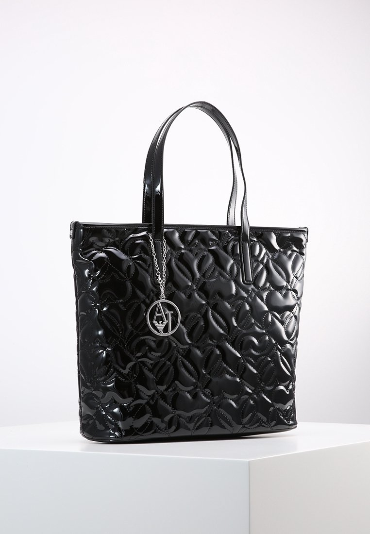Czarna torba na ramię wykonana z błyszczącego materiału, z pikowanym wzorem, płaskimi rączkami oraz okrągłym srebrnym zawieszką z inicjałami.