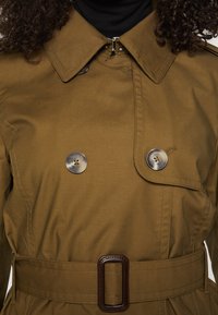 Gros plan d’un trench marron avec de gros boutons, une taille ceinturée et un col sur un pull à col roulé noir.