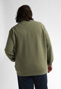 Suéter verde de punto con un patrón texturizado de espiga, cuello alto y puños acanalados. Corte relajado, detalles de costura visibles.
