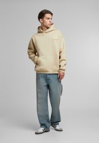 Beige hættetrøje med en kængurulomme og subtil tekst i orange. Matchet med lyseblå jeans og sort-hvide sneakers.