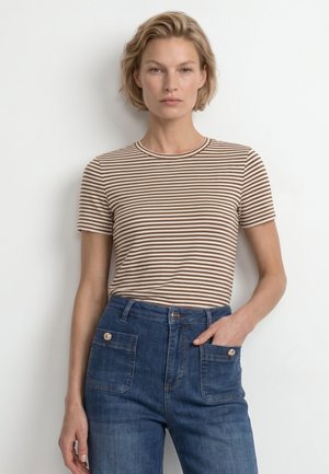 Femme aux cheveux blonds courts portant un t-shirt beige et blanc rayé et un jean bleu taille haute avec de grandes poches à l'avant.