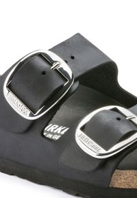 Des sandales Birkenstock noires présentant deux sangles réglables avec des boucles en argent, une semelle intérieure en liège, une finition lisse et une semelle en caoutchouc texturée.