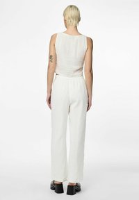 Witte, gestructureerde tanktop gecombineerd met losse, witte broeken. Achteraanzicht toont een schoon ontwerp en een hoge taille. Zwarte hakken.