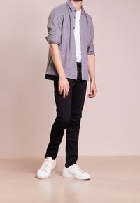 Mand, der står iført en grå skjorte over en hvid t-shirt, sorte bukser og hvide sneakers mod en ensfarvet beige baggrund.