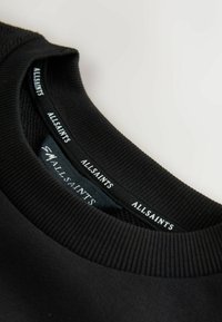 Svart sweatshirt med ribbad textur vid nacken. Har två etiketter: "ALLSAINTS" i vitt och en mindre tagg med ytterligare varumärkesdetaljer.