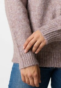 Lichtroze gebreide trui met een geribbelde zoom en manchetten, met een zachte, gestructureerde afwerking. Hand rustend op de mouw voor detail.