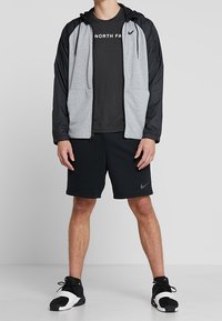 Šedá a černá zipová mikina s kapucí na šňůrku, spárovaná s černým tričkem a černými kraťasy, na oblečení a obuvi jsou zobrazeny loga Nike.